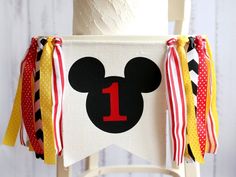 a mickey mouse 1st birthday banner hanging from a chair with red, white and yellow ribbons