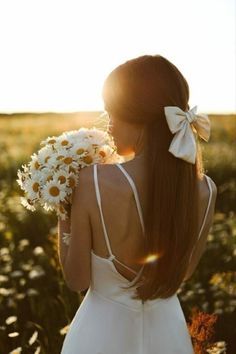 a woman in a white dress holding flowers and looking at the sun through her hair