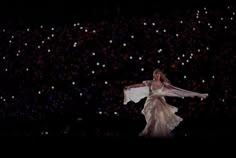 a woman in a white dress is dancing with her arms spread out and lights are behind her