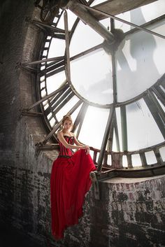 a woman in a long red dress is sitting on a large clock tower with her hands behind her back