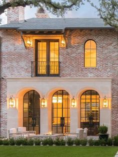 a large brick house with lots of windows and lights on the front door, surrounded by lush green grass