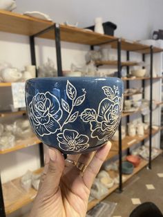 a hand holding up a blue bowl with white flowers on it in front of shelves