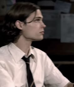 a man wearing glasses and a tie sitting in front of a computer desk with papers on the wall behind him