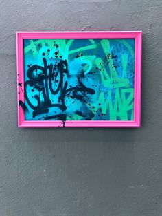 a pink frame with black and blue graffiti on it hanging on the wall next to a gray wall