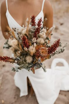 a woman in white dress holding a bouquet of dried flowers and foliage on the beach