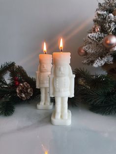 two white candles sitting on top of a table next to a christmas tree and pine cones