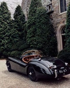 an old fashioned sports car parked in front of a building with trees and bushes around it