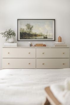 a white bed sitting next to a painting on top of a wooden dresser in a bedroom