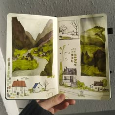 a hand holding an open book with drawings on the pages and houses in the background