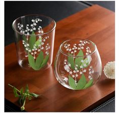 two clear glass vases with flowers painted on them sitting on a wooden table next to each other