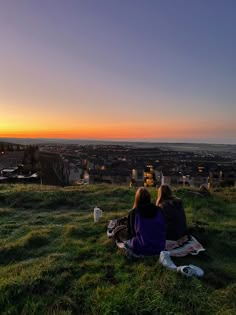 two people sitting on top of a grass covered hill watching the sun set over town