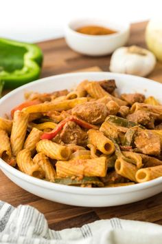 a white bowl filled with pasta and meat on top of a wooden cutting board next to green peppers