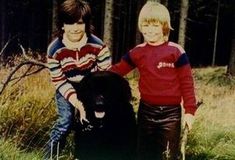 two young boys standing next to each other in the grass with a black bear behind them