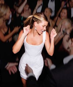 a woman in a white dress is dancing with her hands up and people behind her