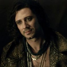 a man with long hair wearing a snake skin jacket and beaded necklaces, looking at the camera