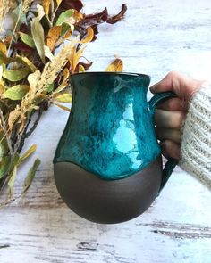 a person holding a green and brown coffee mug next to some dried leaves on a white wooden table