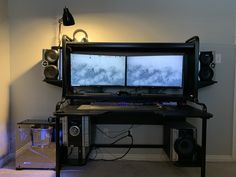 two computer monitors sitting on top of a black desk in front of a white wall