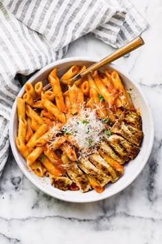 a white bowl filled with pasta and chicken covered in parmesan cheese on top of a marble table