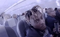 a man with dreadlocks sitting in an airplane looking at his cell phone and taking a selfie