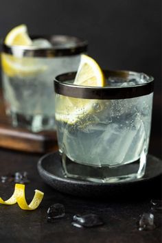 two glasses filled with ice and lemons on top of a black plate next to a cutting board