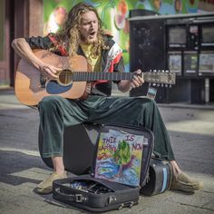 a man sitting on the ground with a guitar in his hand and an open suitcase behind him