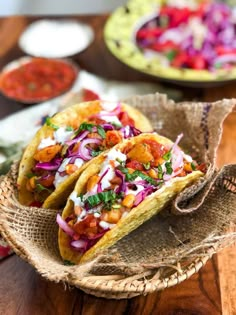 two tacos are sitting in a basket on a wooden table with other food items