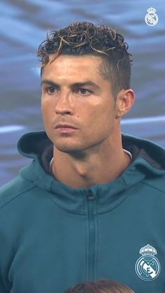 a man with curly hair standing in front of a blue background wearing a green hoodie