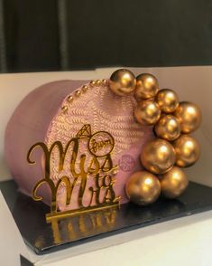 the cake is decorated with gold balls and a pink frosted hat on it's stand