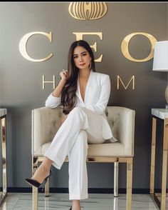 a woman sitting on a chair in front of a wall with gold letters and a white suit