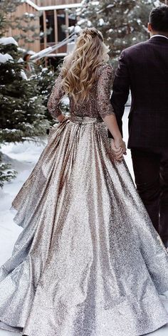 a man and woman are walking in the snow holding hands, dressed in silver dresses