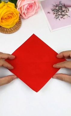 two hands holding a piece of red paper on top of a table with flowers and pictures