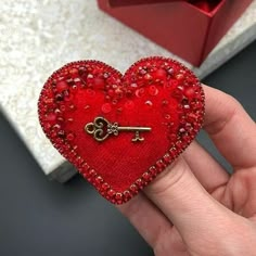 a hand holding a red heart shaped brooch with a golden key on it's side