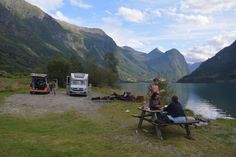 two people are sitting at a picnic table near the water with mountains in the background