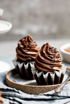 three chocolate cupcakes sitting on top of a wooden plate next to a bowl of frosting