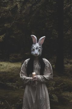 a person wearing a rabbit mask and holding a candle in the woods with trees behind them