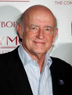 an older man wearing a blue shirt and black blazer smiles at the camera while standing in front of a red carpeted wall