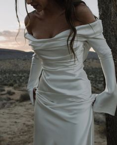 a woman in a white dress standing next to a tree with her hands on her hips