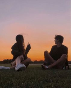 two people sitting in the grass at sunset, one holding a cell phone and the other looking up