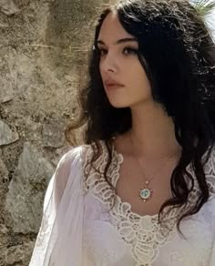 a woman wearing a white shirt and necklace standing in front of a stone wall with her hand on her hip