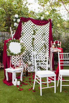 an outdoor wedding ceremony with white chairs and red drapes on the back wall, decorated with flowers and greenery