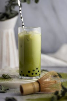 a glass filled with green liquid next to a white vase and brush on a table