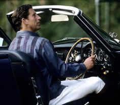 a man sitting in the driver's seat of a classic car with his hand on the steering wheel