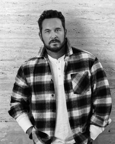 a man standing in front of a brick wall wearing a plaid shirt and jeans with his hands in his pockets