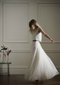 a woman in a long white skirt and crop top is standing by a table with flowers