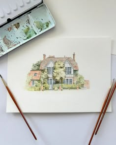 a watercolor painting of a house on paper next to two paintbrushes and an artist's palette