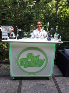 a woman standing behind an outdoor bar with bottles on the counter and glasses on it