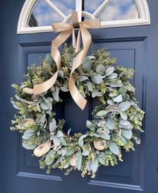 a wreath is hanging on the front door with a gold ribbon and some greenery