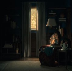 a woman sitting on a chair in a dark room with a teapot and lamp