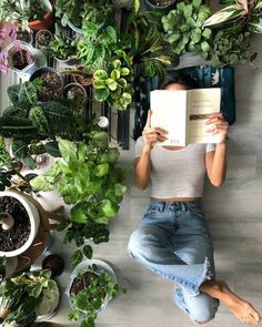 a woman sitting on the floor reading a book surrounded by plants and potted plants