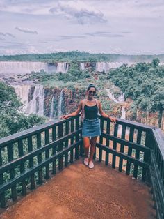 a woman standing at the edge of a bridge overlooking a waterfall and trees in the background